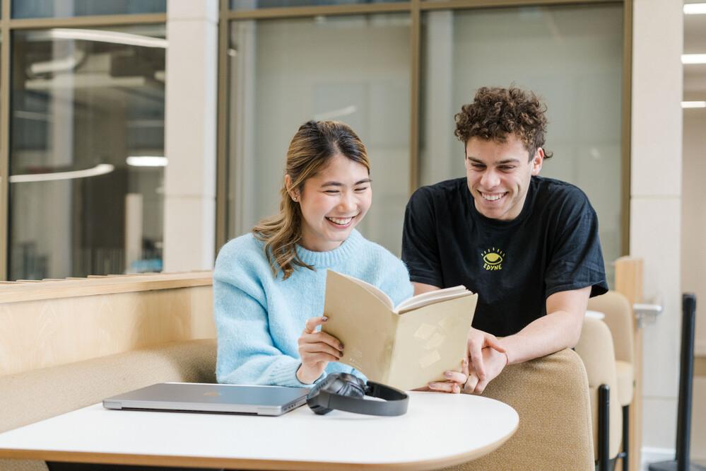 Two students are reading a book at a desk. They are smiling.